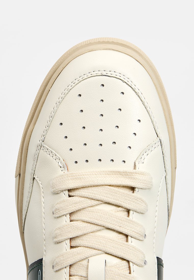 Zapatilla con parte superior de cuero sintético color crema, puntera redonda, detalles perforados, cordones beige y suela de goma color marrón claro.