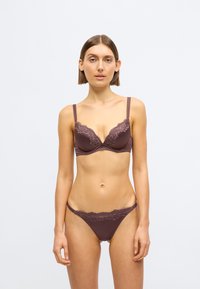 Rudos spalvos liemenėlės ir kelnaičių komplektas su nėrinių apdaila. Liemenėlė turi lankelius ir reguliuojamas petnešėles, o kelnaitės pasižymi panašiu nėrinių dizainu.