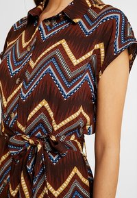 Robe chemise marron à manches courtes avec un motif en zigzag. Présente des accents bleus, jaunes et noirs, des boutons de fermeture et une taille nouée.