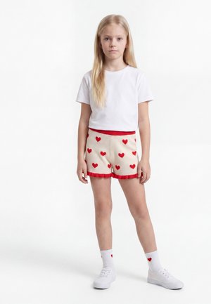 Jeune fille aux longs cheveux blonds portant un T-shirt blanc, un short crème avec des cœurs rouges, des chaussettes blanches avec des cœurs rouges et des baskets blanches, debout.