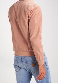 Jasnoróżowa kurtka bomberka wykonana z gładkiego materiału, z prążkowymi mankietami i dolnym ściągaczem, z czystym tyłem, zestawiona z jasnoniebieskimi dżinsami.