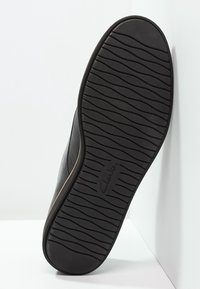 Semelle de chaussure en caoutchouc noir avec un motif de bande de roulement texturé en forme de vagues. Présente un bord incurvé et un logo de marque embossé. Surface lisse sans marquages visibles.