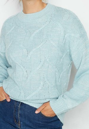 Lyseblå strikket sweater med et kabelmønster, båret med mørkeblå jeans, hænderne i lommerne, beskåret midt på torsoen.