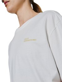 Zbliżenie osoby noszącej białą koszulę z żółtym tekstem "HALO 196819721981" na piersi, włosy związane w kucyk.