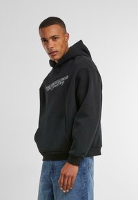 Sort hættepullover lavet af blødt stof, med en frontlomme og en hætte med snørelukning. Teksten på fronten lyder: "GØR DIG SELV STOLTE."