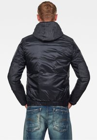 Man med kort brunt hår som bär en svart huvjacka med vadderade armbågar och blå jeans i en ljusare nyans, stående med händerna i fickorna.