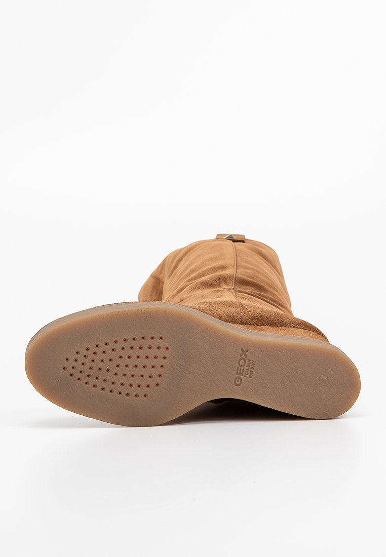 Bottine en daim marron avec un bout arrondi et une semelle en caoutchouc flexible. La semelle présente de petites perforations et un logo sur le côté.