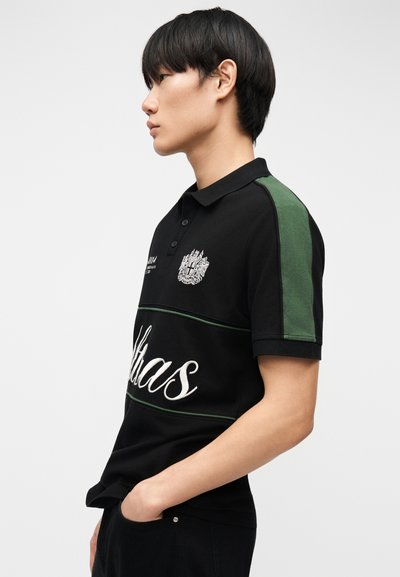 Νεαρός άνδρας με μαύρο πόλο με πράσινη γραμμή στον ώμο και λευκό κεντημένο κείμενο, στέκεται σε προφίλ με τα χέρια στις τσέπες.