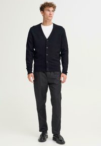 Svart strikket cardigan med V-hals og seks knapper, parret med svarte bukser og svarte sko. Modellen står med hendene på sidene.
