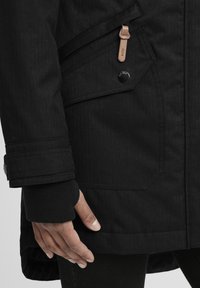 Veste imperméable noire avec une finition texturée, dotée d'une poche latérale, de détails en bouton et de poignets en tricot. Main visible au niveau de l'ourlet.