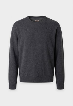 Homme debout portant un pull gris foncé sur une chemise rayée, un pantalon beige et des chaussures habillées noires sur un fond uni clair.