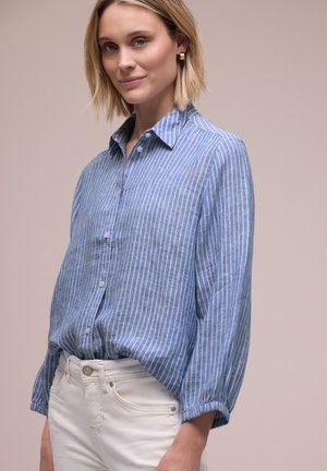 Frau mit blondem Haar, die goldene Creolenohrringe, ein blau-weiß gestreiftes Hemd mit Knöpfen und weiße Hosen trägt, steht vor einem neutralen Hintergrund.