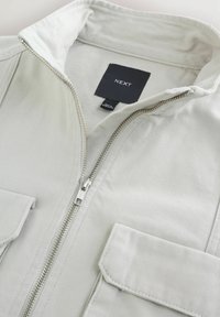 Jachetă gri deschis cu un fermoar frontal parțial deschis, care arată gulerul și eticheta neagră cu brandul „NEXT” în interiorul zonei gâtului.