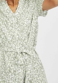 Femme portant une robe à manches courtes boutonnée à motifs vert clair et blanc, avec une ceinture nouée, devant un fond uni.