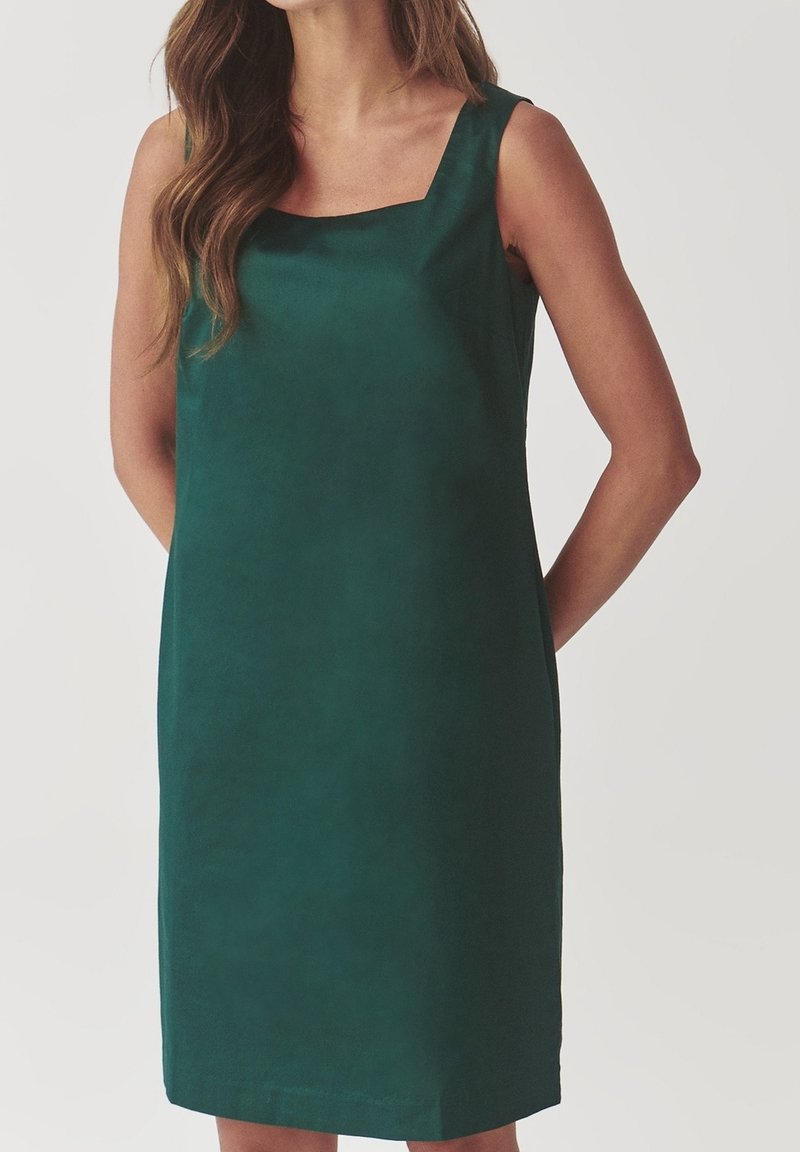 Femme portant une robe sans manches vert foncé longueur genou avec un décolleté carré, debout les mains dans le dos.