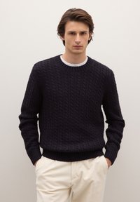 Giovane uomo con le mani nelle tasche, che indossa un maglione nero a trecce sopra una camicia bianca e pantaloni crema, davanti a un muro chiaro e uniforme.