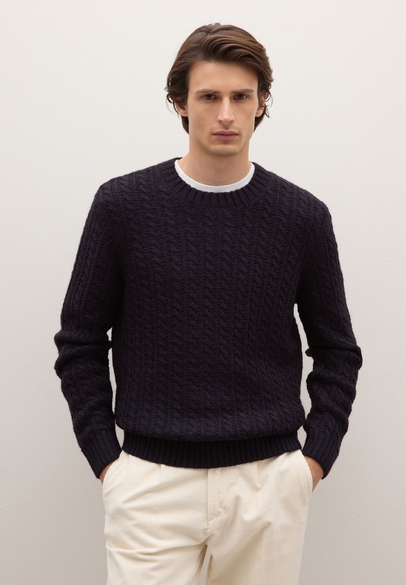 Giovane uomo con le mani nelle tasche, che indossa un maglione nero a trecce sopra una camicia bianca e pantaloni crema, davanti a un muro chiaro e uniforme.