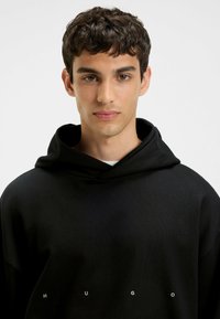 Black hoodie made from textured fabric, featuring a drawstring hood and the word "HUGO" in white lettering across the chest.