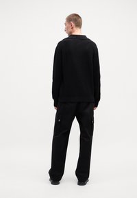 Schwarzer Strickpullover mit rundem Ausschnitt, kombiniert mit schwarzen Cargo-Hosen mit seitlichen Taschen und Druckknopf-Details.