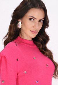 Helles pinkes Strickpullover mit hohem Ausschnitt, geschmückt mit bunten, gem-artigen Verzierungen. Das Model trägt große Tropfenohrringe.