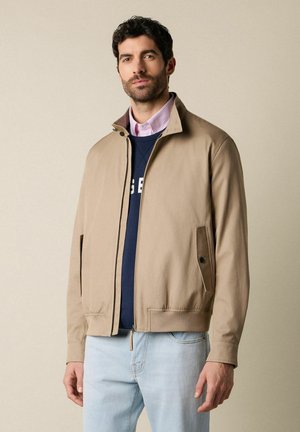 Mężczyzna w beżowej kurtce na zamek, granatowej bluzie, różowej koszuli z kołnierzykiem i jasno-niebieskich dżinsach, stojący na tle jednolitego beżowego tła.