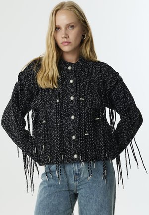 Giacca texturizzata in bianco e nero con lungo frange, dotata di grandi bottoni e colletto alto. Abbinata a jeans blu chiaro.