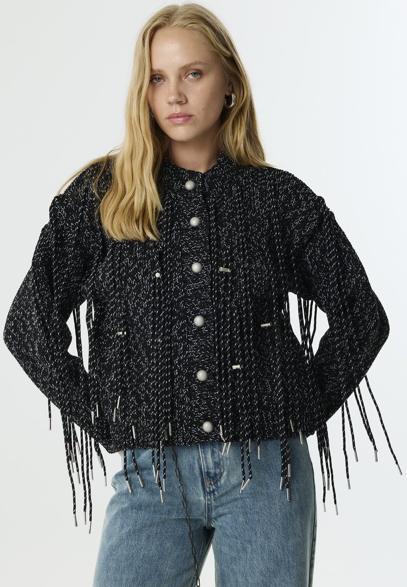 Chaqueta texturizada en blanco y negro con largas borlas, presenta grandes botones y un cuello alto. Combinada con jeans de color azul claro.
