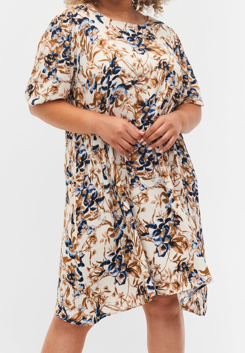 Femme portant une robe crème à manches courtes avec un imprimé floral bleu et marron, tenant le tissu de la robe près de la taille.