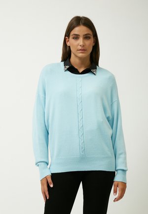 Lichtblauwe oversized trui met kabelbrei-details in het midden en geribde manchetten, gedragen over een zwarte blouse met kraag en versieringen.