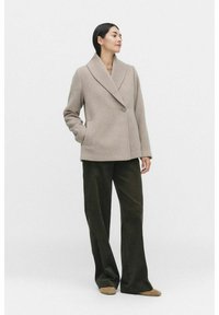 Beige wool coat with a shawl collar and single button closure, worn over olive wide-leg pants. Smooth texture, minimalist design.