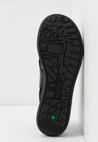 Semelle d'une chaussure Timberland noire avec motif de bande de roulement géométrique, logo hexagonal vert près du talon, affichée droite sur fond blanc.