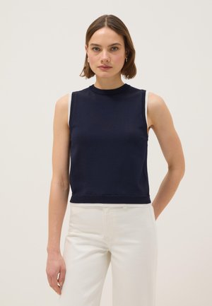 Vrouw met kort bruin haar draagt een mouwloos marineblauw topje en witte broek, staand tegen een effen lichte achtergrond.