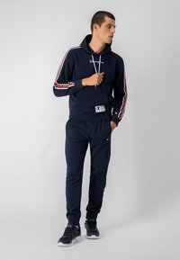 Sudadera azul marino con cordones blancos y logo, con rayas rojas y blancas en las mangas. Combinada con pantalones de chándal a juego y zapatillas oscuras.