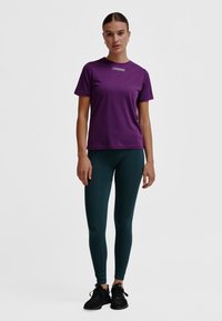 Lila sport-t-shirt med korta ärmar och rund hals, kombinerad med mörkgröna ribbade leggings. Svarta sportskor fullbordar outfiten.
