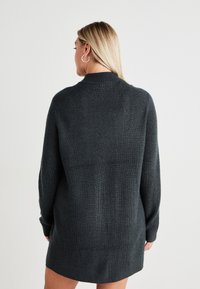 Dark gray, ribbed knit sweater with long sleeves and a high neck, featuring a relaxed fit and mid-thigh length. No prominent patterns.