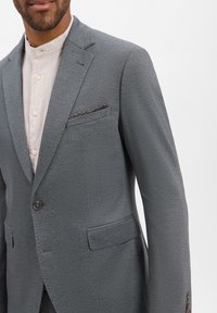 Grijs op maat gemaakt pak van textuurstof, met een enkele knoopsluiting en twee voorzakken. Voorzien van een borstzak die is geaccentueerd met een gestreepte zakdoek.