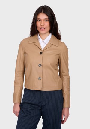 Žena s dlouhými tmavými vlasy, která má na sobě hnědou koženou bundu na knoflíky přes bílou košili a tmavé kalhoty s proužky, stojící před jednobarevným pozadím.