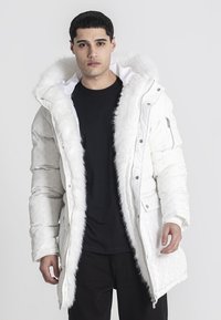 Bijela puffer jakna s umjetnim krznom, džepovima na zip, i uzorkovanom školjkom. Model nosi crnu majicu ispod.