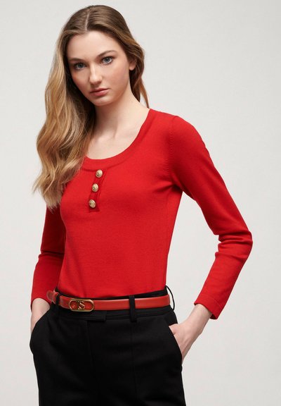 Red long-sleeve top with a scoop neckline and three decorative gold buttons at the chest, paired with high-waisted black trousers and a red belt.