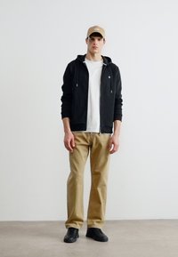 Jeune homme debout portant une casquette de baseball beige, un sweat à capuche noir zippé, un t-shirt blanc, un pantalon beige et des baskets noires devant un mur blanc uni.