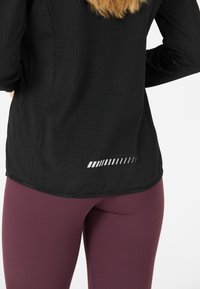 Schwarzes langärmliges Sportoberteil aus strukturiertem Stoff, mit einem reflektierenden horizontalen Streifendesign nahe dem Saum, kombiniert mit lila Leggings.