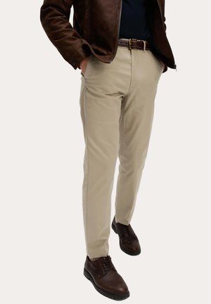 Pantalones beige con textura suave, de corte recto, con bolsillos laterales y cinturón marrón oscuro. Combinados con zapatos marrón oscuro con cordones.