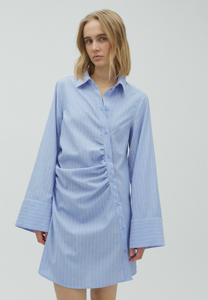 Jeune femme portant une robe boutonnée bleu clair à manches longues avec des rayures blanches verticales et de larges poignets aux manches.