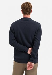 Marineblauwe ribgebreide trui met lange mouwen en ronde halslijn, met een getextureerde stof en smalle manchetten, gedragen met beige broek.