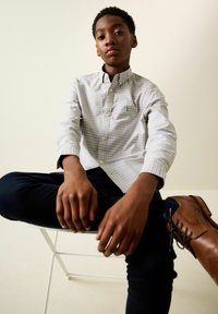 Világos tónusú kockás gombos ing, sötét nadrággal és barna bőr cipővel párosítva. Figyelemre méltó részletek közé tartozik egy mellzseb és mandzsetták.