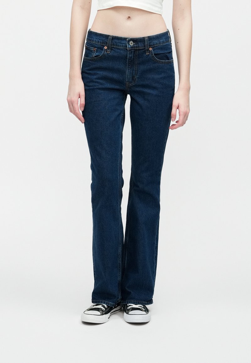 Kobieta w ciemnoniebieskich prostych spodniach dżinsowych i czarnych niskich sneakersach, stojąca na tle białego tła.