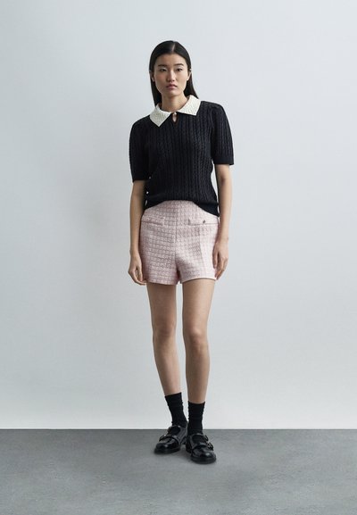 Femme portant un haut en maille noir à manches courtes avec un col blanc, un short rose clair texturé, des chaussettes noires et des mocassins noirs, debout sur un sol gris.