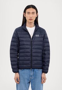 Navy puffer jacket with horizontal quilted design, high collar, and full zipper. Features EA7 logo on the left chest. Worn over a white t-shirt.