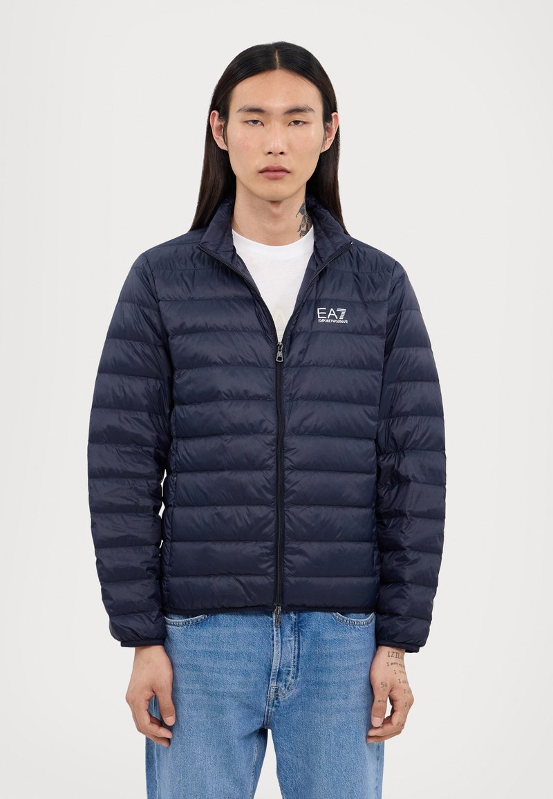 Navy puffer jacket with horizontal quilted design, high collar, and full zipper. Features EA7 logo on the left chest. Worn over a white t-shirt.