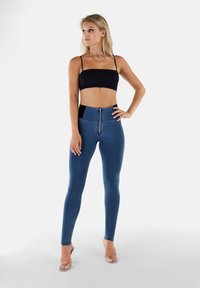 Leggings in denim blu alti in vita con zip frontale, cintura nera e pannelli laterali, abbinati a un top nero corto.